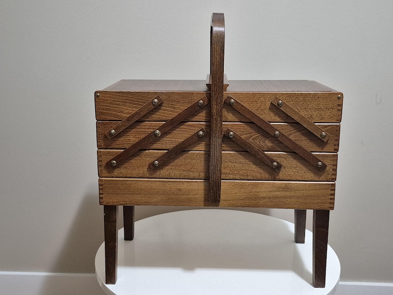 May include: A vintage wooden sewing box with a handle, featuring a multi-tiered design that expands outwards. The box is made of light brown wood, with brass-colored hardware and sits on four short legs. The handle is arched and made of the same wood.