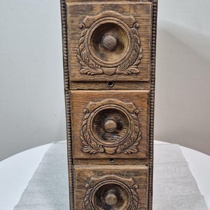 May include: A vertical wooden storage unit featuring three stacked drawers. Each drawer front displays a circular carved design with a central knob and leaf-like patterns. The wood has a medium brown finish with darker stained accents.