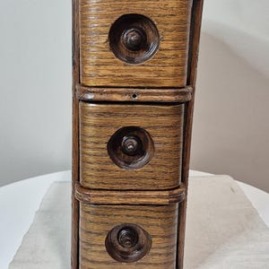May include: A vintage wooden three-drawer storage unit. The drawers are rectangular with a circular design in the centre and a dark brown knob. The unit is made of light brown wood with a visible wood grain.