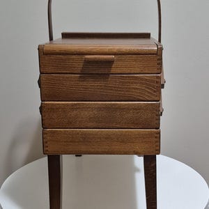 May include: A vintage wooden sewing box with a handle, featuring three drawers and a top compartment. The box is supported by four legs and has a dark brown finish. The handle is curved and attached to the top of the box.
