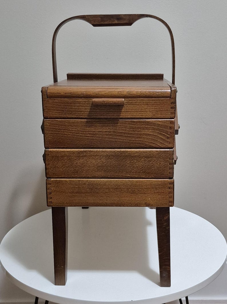 May include: A vintage wooden sewing box with a handle, featuring three drawers and a top compartment. The box is supported by four legs and has a dark brown finish. The handle is curved and attached to the top of the box.