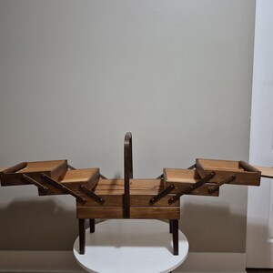 May include: A vintage wooden sewing box with multiple compartments and a handle. The box is made of light brown wood and has a tiered design with drawers that open outwards. It sits on a small, round table.