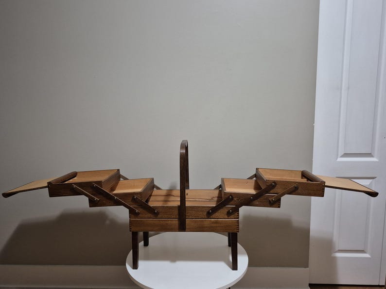 May include: A vintage wooden sewing box with multiple compartments and a handle. The box is made of light brown wood and has a tiered design with drawers that open outwards. It sits on a small, round table.