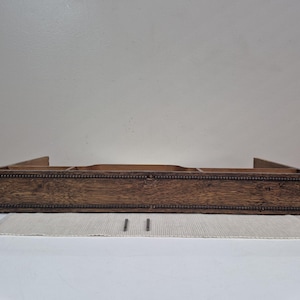 May include: A rectangular wooden shelf with a dark brown wood grain finish and a beaded trim along the top and bottom edges. The shelf features an open interior divided into three sections by two vertical wooden dividers, with two metal pins.