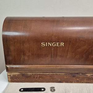 May include: A vintage wooden sewing machine case, branded "SINGER" in gold lettering. The case is a warm brown colour with a curved top and a rectangular base. A black leather strap and metal hardware are visible.
