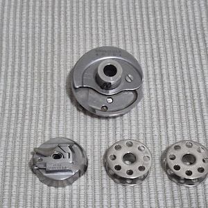 May include: A collection of vintage Singer sewing machine parts. The image shows a silver-colored bobbin case, a bobbin holder, and two bobbins. The parts are arranged on a light gray textured surface.