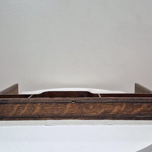 May include: A rectangular dark brown wooden desk organiser featuring a long central compartment flanked by two smaller side sections. The front panel displays a prominent wood grain pattern and is accented with a horizontal beaded trim detail.