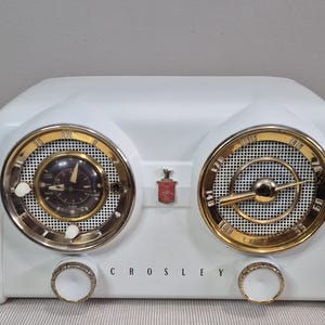May include: A white Crosley radio with a dual-dial design featuring gold-toned circular bezels, a clock on the left, a radio tuner on the right, two white control knobs at the bottom, and a small red emblem centred between the two main dials.