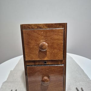 May include: A small, two-drawer wooden cabinet with a warm, brown finish. Each drawer has a round, wooden knob. The cabinet is rectangular and appears to be vintage or antique. Two screws are visible near the cabinet.
