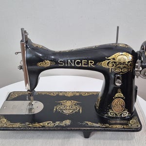 May include: A vintage black Singer sewing machine featuring intricate gold filigree decals across the body and base. The machine includes a prominent silver handwheel on the right side, a needle bar, and a metal throat plate with visible text.