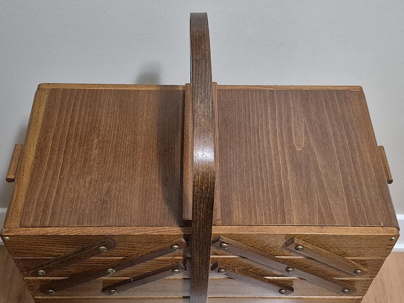 May include: A wooden sewing box with a dark brown finish. The box has a handle and a multi-tiered design with pull-out trays. The box is made of wood and has a classic, vintage aesthetic.