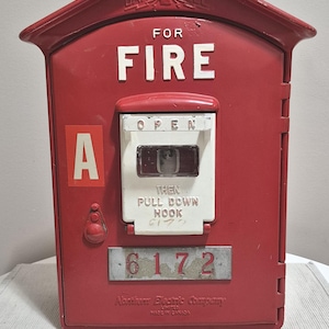 Peut inclure: Un ancien boîtier d'alarme incendie rouge avec le mot "FIRE" en lettres blanches. Le boîtier a une porte à charnière avec les mots "OPEN" et "THEN PULL DOWN HOOK". En dessous de la porte se trouve le numéro "6172". Deux clés sont en bas.