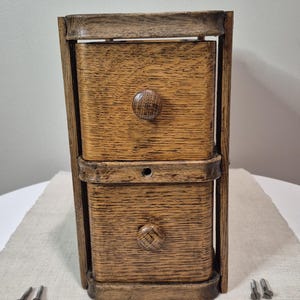 May include: A small, two-drawer wooden cabinet with a natural wood grain finish. The cabinet has a dark brown frame and round wooden knobs. The drawers are rectangular and appear to be made of oak. Two small screws are visible near the base.
