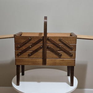 May include: A wooden sewing box with a handle and legs. The box has a tiered design with hinged compartments that open outwards. The wood has a warm, medium brown tone, and the box is set on a white surface.