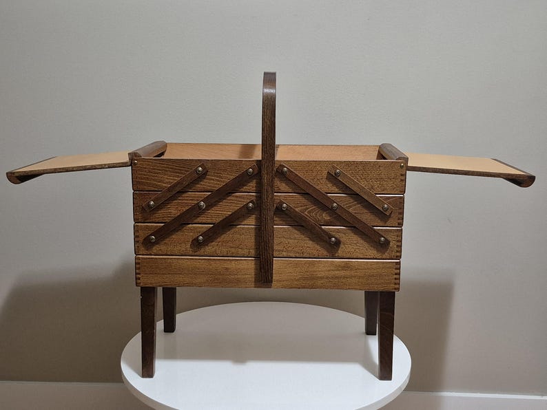 May include: A wooden sewing box with a handle and legs. The box has a tiered design with hinged compartments that open outwards. The wood has a warm, medium brown tone, and the box is set on a white surface.