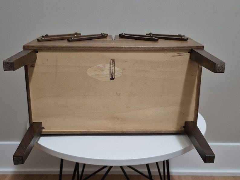 May include: A wooden, rectangular coffee table with dark brown legs and a light brown tabletop. The table is upside down, revealing the underside with a light wood finish and a small, oval-shaped sticker. The table is on a white table.
