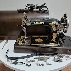 May include: Vintage Singer sewing machine, black with gold accents, and a wooden carrying case. Accessories include a screwdriver, presser feet, and a thimble. The machine is on a white table.