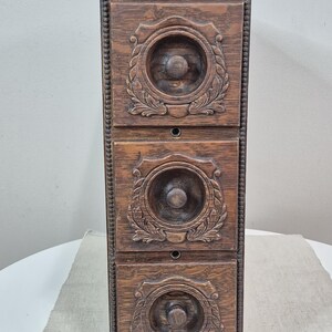 May include: A vertical wooden storage unit featuring three stacked drawers. Each drawer front is decorated with a circular carved motif surrounded by leaf patterns and a central knob. The frame has a beaded edge detail along the vertical sides.