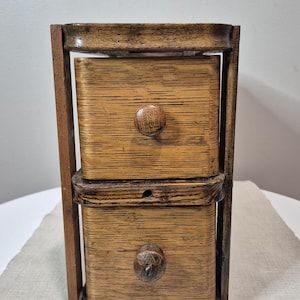 Peut inclure: Petit meuble en bois à deux tiroirs, avec un cadre marron foncé et des tiroirs marron clair. Chaque tiroir est doté d'un bouton rond en bois. Le meuble est en bois avec un grain visible et est probablement vintage ou antique.