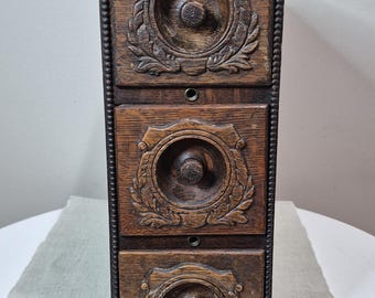 May include: A vertical wooden storage unit featuring three stacked drawers. Each drawer front displays a circular carved relief pattern with a central knob. The edges of the unit are decorated with a beaded trim detail in a dark brown wood finish.