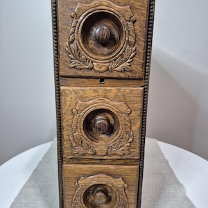 May include: A vertical wooden storage unit featuring three stacked drawers. Each drawer front displays a circular recessed centre with a central knob, surrounded by carved leaf-like decorative patterns. The frame has a beaded edge detail.