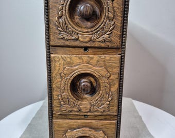 May include: A vertical wooden storage unit featuring three stacked drawers. Each drawer front displays a circular recessed center with a central knob, surrounded by carved leaf-like decorative patterns. The frame has a beaded edge detail.