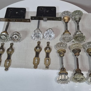 May include: A collection of antique door hardware, including glass doorknobs, brass keyhole covers, and metal latches. The doorknobs have a clear, faceted design. The hardware is arranged on a neutral-colored fabric.