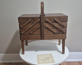 May include: A vintage wooden sewing box with a handle, featuring multiple compartments that expand outwards. The box is a warm brown color, with brass-colored hardware. It stands on four short legs and sits on a white table.