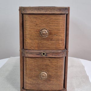Peut inclure: Petit meuble en bois à deux tiroirs avec une finition naturelle. Les tiroirs ont des boutons ronds de couleur laiton. Le meuble est rectangulaire avec un cadre marron foncé, probablement vintage ou antique.