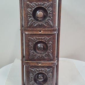 May include: A dark brown, three-drawer wooden cabinet with ornate floral carvings on each drawer front. The drawers have round knobs and are framed by a vertical wooden structure. The cabinet is likely vintage or antique.