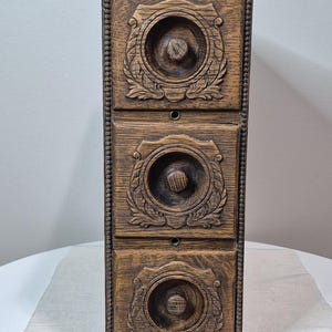 May include: A vertical wooden storage unit featuring three stacked drawers. Each drawer front is decorated with a circular carved relief pattern and a central wooden knob. The edges of the unit are detailed with a beaded trim design.