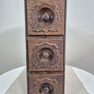 May include: A vertical wooden storage unit featuring three stacked drawers. Each drawer front is decorated with a circular carved relief pattern and a central wooden knob. The frame of the unit is accented with a beaded edge detail along the sides.
