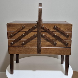 May include: A vintage wooden sewing box with a handle and four pull-out drawers. The box is made of dark wood and has a hinged lid. The drawers are secured with wooden supports and metal studs. The box sits on four short legs.