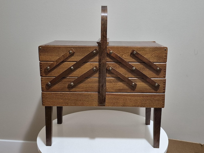 May include: A vintage wooden sewing box with a handle and four pull-out drawers. The box is made of dark wood and has a hinged lid. The drawers are secured with wooden supports and metal studs. The box sits on four short legs.