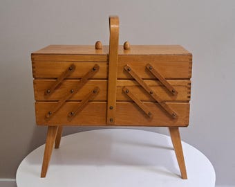 May include: A vintage wooden sewing box with a handle and angled legs. The box is light brown with a hinged lid and multiple compartments. The box has a classic, retro design.