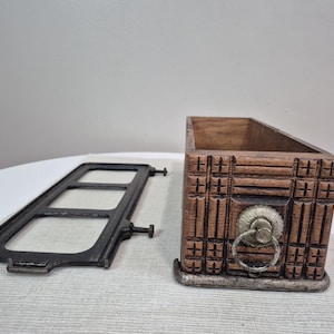 May include: A wooden box with carved geometric patterns and a metal handle. Beside it is a black metal frame with rectangular openings. The box is brown, and the metal elements are dark.