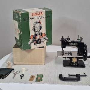 May include: A black Singer Sewhandy sewing machine with its original green and white box featuring an illustration of a person sewing. Included are a metal C-clamp, small scissors, a thimble, a wooden spool, a needle packet, and a black case.