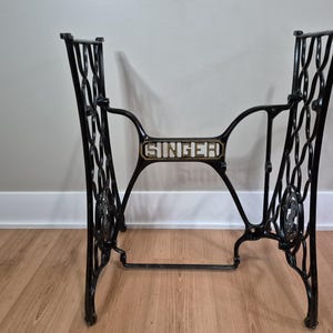 May include: A black cast iron Singer sewing machine base featuring an intricate, openwork scroll design on the side supports. The central horizontal brace displays the word SINGER in raised, light-coloured lettering against a dark background.