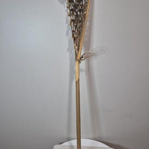 May include: A vintage lacrosse stick with a wooden shaft and a woven net head. The stick has a light brown color with darker brown netting. The stick is standing upright on a white surface.