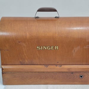 May include: Vintage Singer sewing machine case made of light brown wood. The case has a curved top, a dark brown handle, and the word "SINGER" in gold lettering. The case is rectangular with a small circular opening on the bottom right.