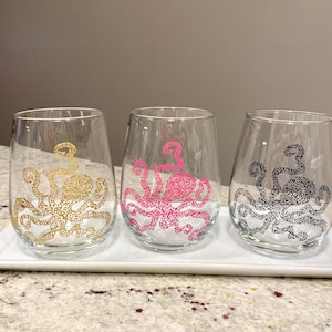 May include: Three stemless wine glasses with octopus designs in gold, pink, and black. The octopus illustrations are detailed and cover most of the glass surface. The glasses are displayed on a white rectangular tray.