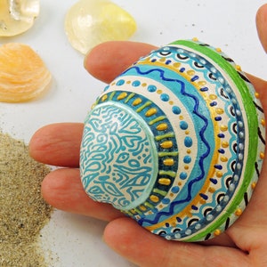 Op de afbeelding: Een handgeschilderde schelp met een geometrisch patroon in blauw, groen en goud. De schelp is versierd met witte stippen en spiralen.