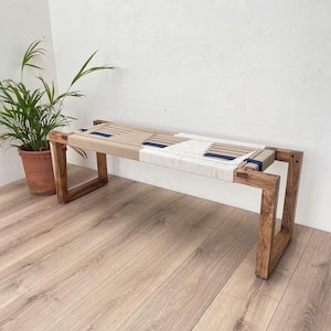 May include: A wooden bench with a woven seat made of natural fibers in shades of beige and white with accents of navy blue. The bench has a simple, rectangular design with a sturdy wooden frame.