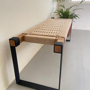 May include: A wooden bench with a woven rope seat and black metal legs. The bench has a natural wood finish and the rope is a light beige color.