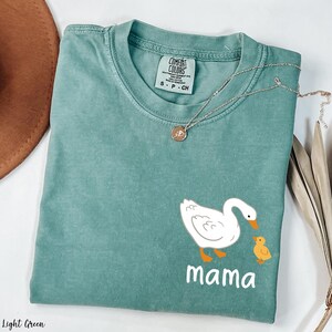 K&ouml;nnte beinhalten: Hellgr&uuml;nes T-Shirt mit einer wei&szlig;en Ente und einem gelben Entenk&uuml;ken. Der Text "mama" ist unter den Enten gedruckt.