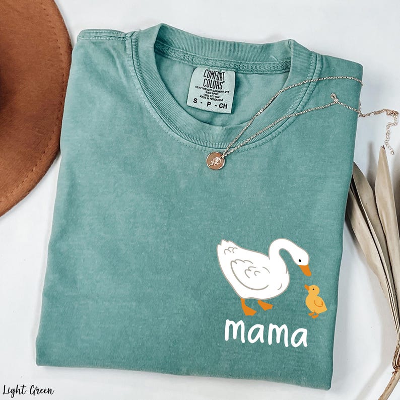 K&ouml;nnte beinhalten: Hellgr&uuml;nes T-Shirt mit einer wei&szlig;en Ente und einem gelben Entenk&uuml;ken. Der Text "mama" ist unter den Enten gedruckt.