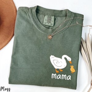 K&ouml;nnte beinhalten: Ein gr&uuml;nes T-Shirt mit einer wei&szlig;en Ente und einem gelben Entenk&uuml;ken. Der Text "mama" ist unter den Enten gedruckt.