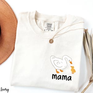K&ouml;nnte beinhalten: Ein wei&szlig;es T-Shirt mit einem Aufdruck eines wei&szlig;en Entenpaares, bestehend aus einer Ente und einem Entenk&uuml;ken. Unter den Enten steht der Text "mama".
