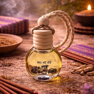 May include: A clear glass aromatherapy bottle with a wooden cap and a rope loop. The bottle contains a yellow liquid and dark stones, with the text "NAG ME NOT - HEMATITE" on the front. The background includes incense and a lit candle.