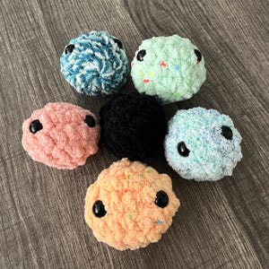 May include: Six colorful, crocheted plush balls with black button eyes. The balls are in shades of blue, green, pink, black, orange, and light blue. The plush toys are arranged on a wooden surface.